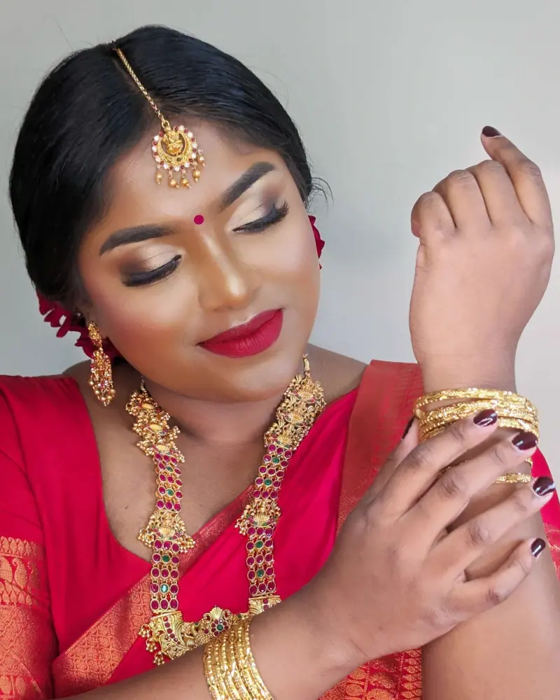 Arthy en saree rouge, lèvres rouges, parure dorée et chignon, main posée à la joue.