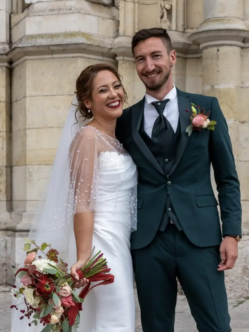 Clémence et son marié devant une façade de pierre, cape de tulle perlée et bouquet rouge et rose.