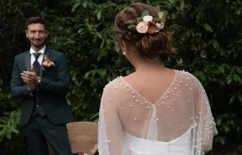 Le marié découvre Clémence de dos, chignon orné de fleurs roses et cape perlée.