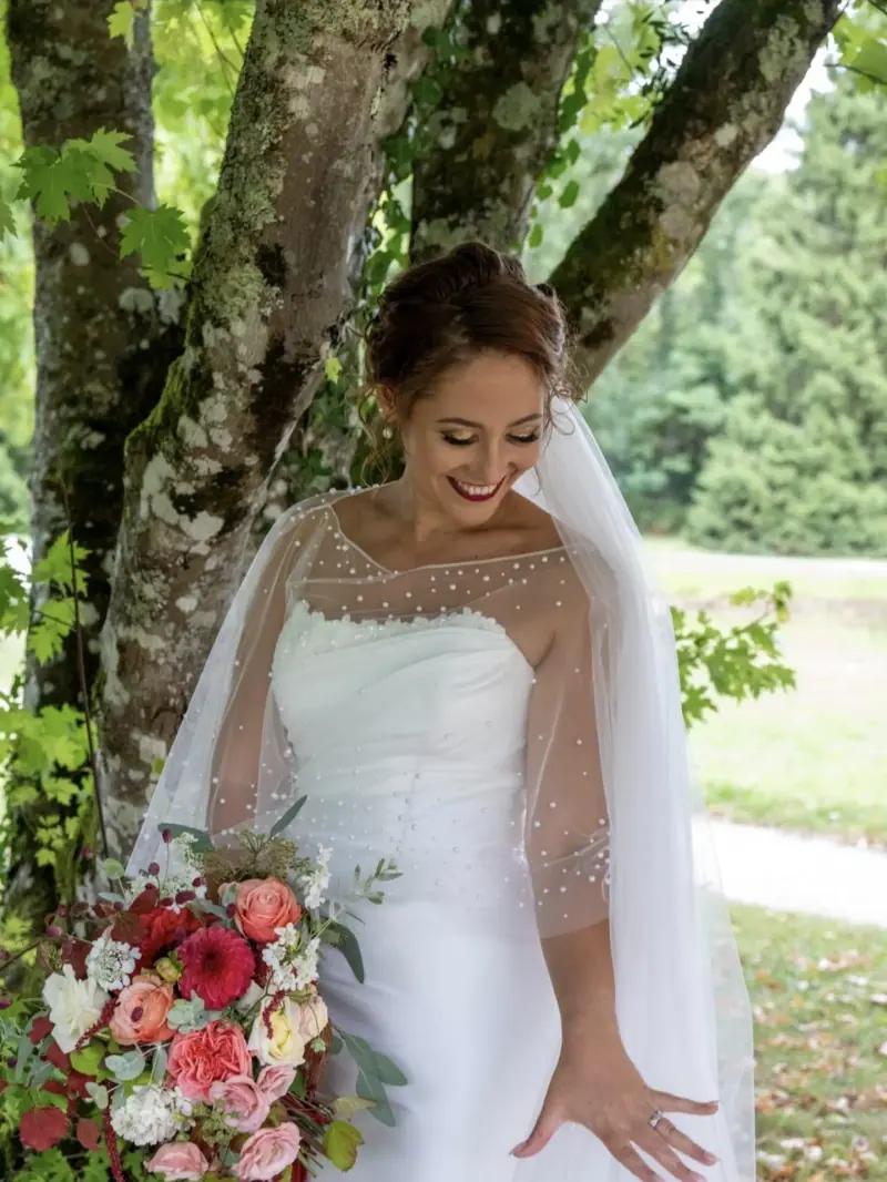 Clémence sous un arbre, voile et bouquet rouge et rose, regard baissé.
