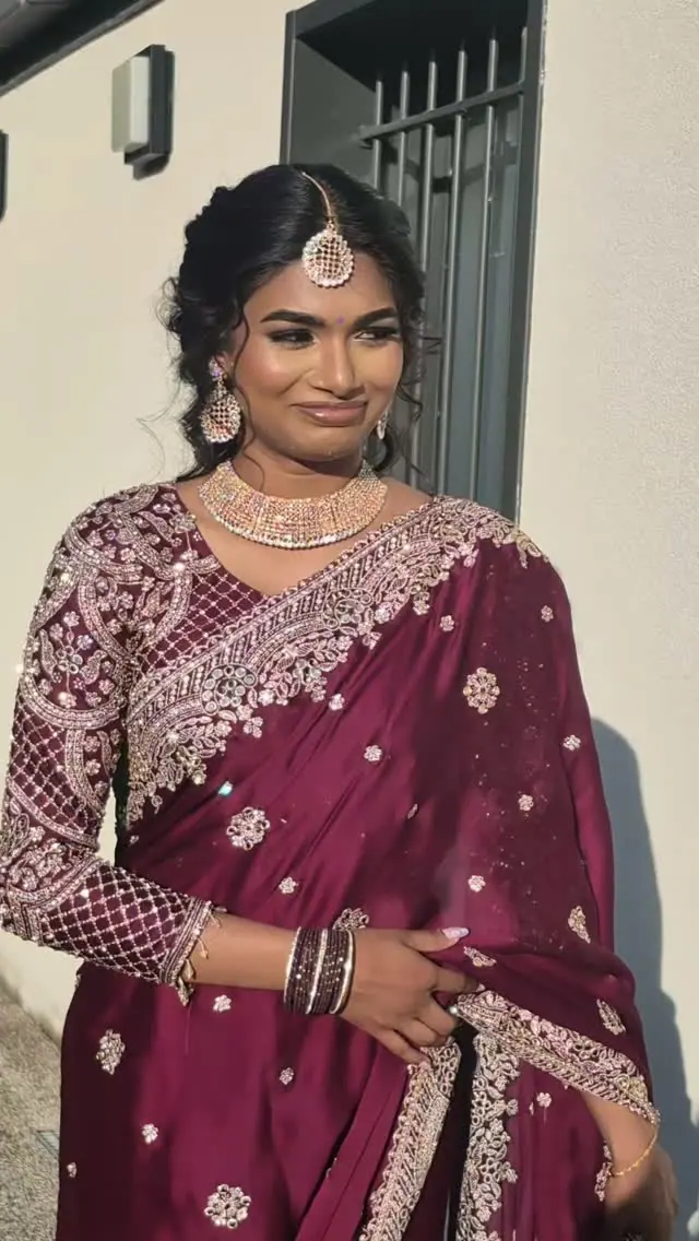 Jeune femme en saree bordeaux brodé, maang tikka et longs cheveux bouclés, souriante en lumière du soir.