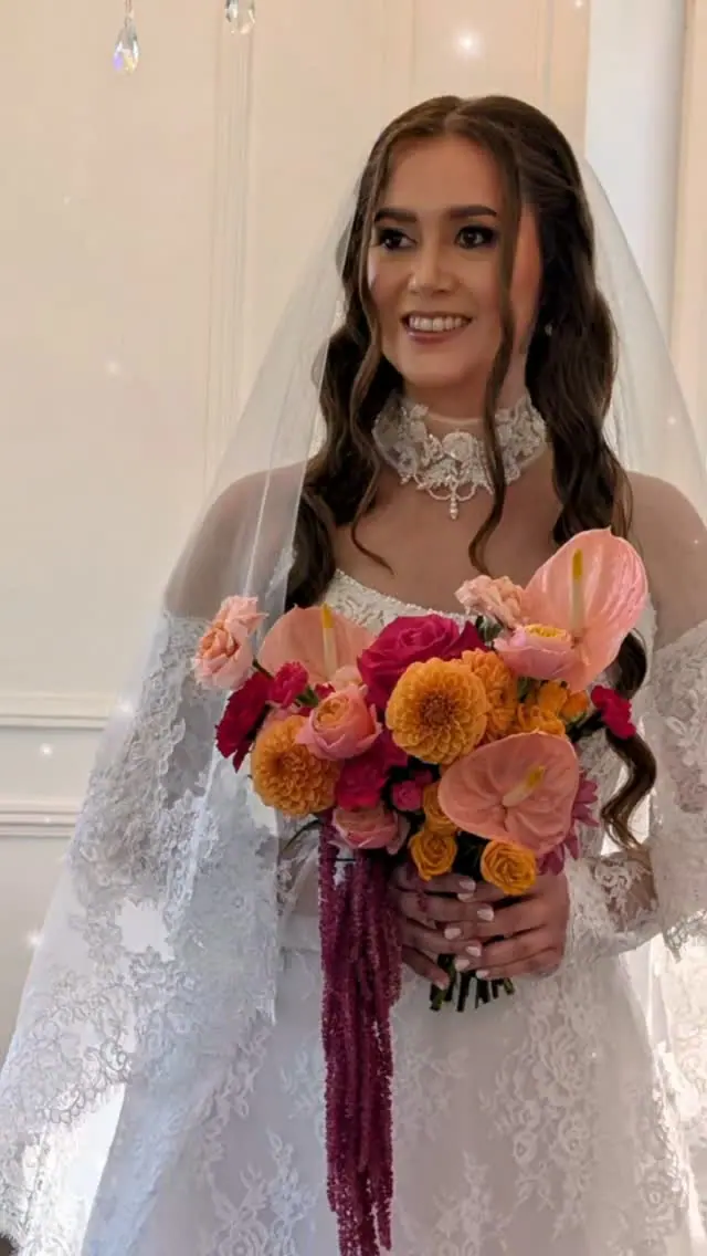 Mariée souriante en robe de dentelle et voile, cheveux longs ondulés, tenant un bouquet orange et rose.