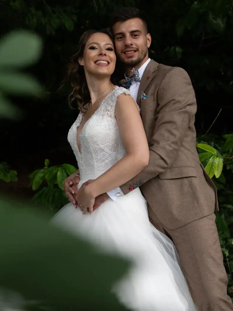 Amandine enlacée par son marié dans un jardin, robe de dentelle et coiffure semi-attachée.