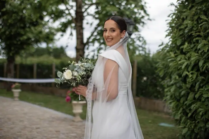 Vanessa de dos dans un jardin, cape de tulle et bouquet blanc.