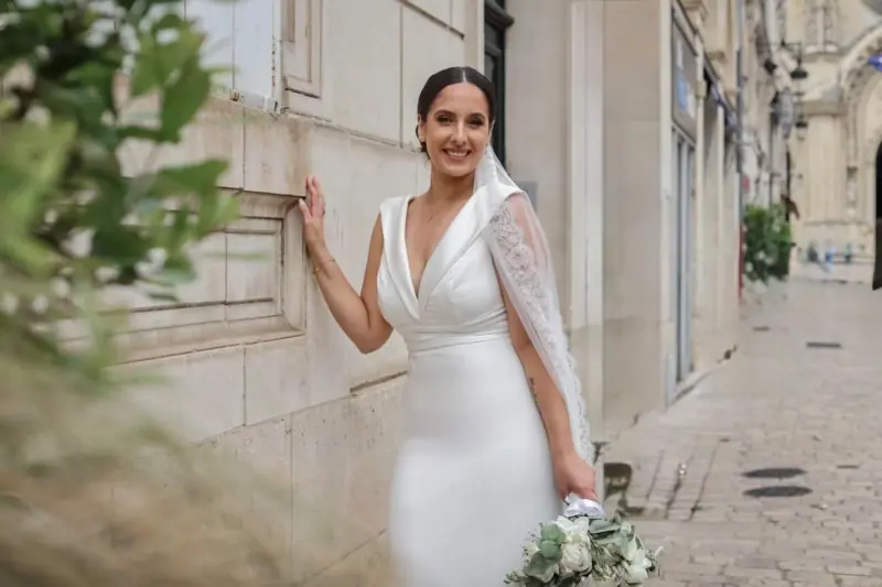 Vanessa souriante dans une rue pavée, cape de dentelle et bouquet blanc.