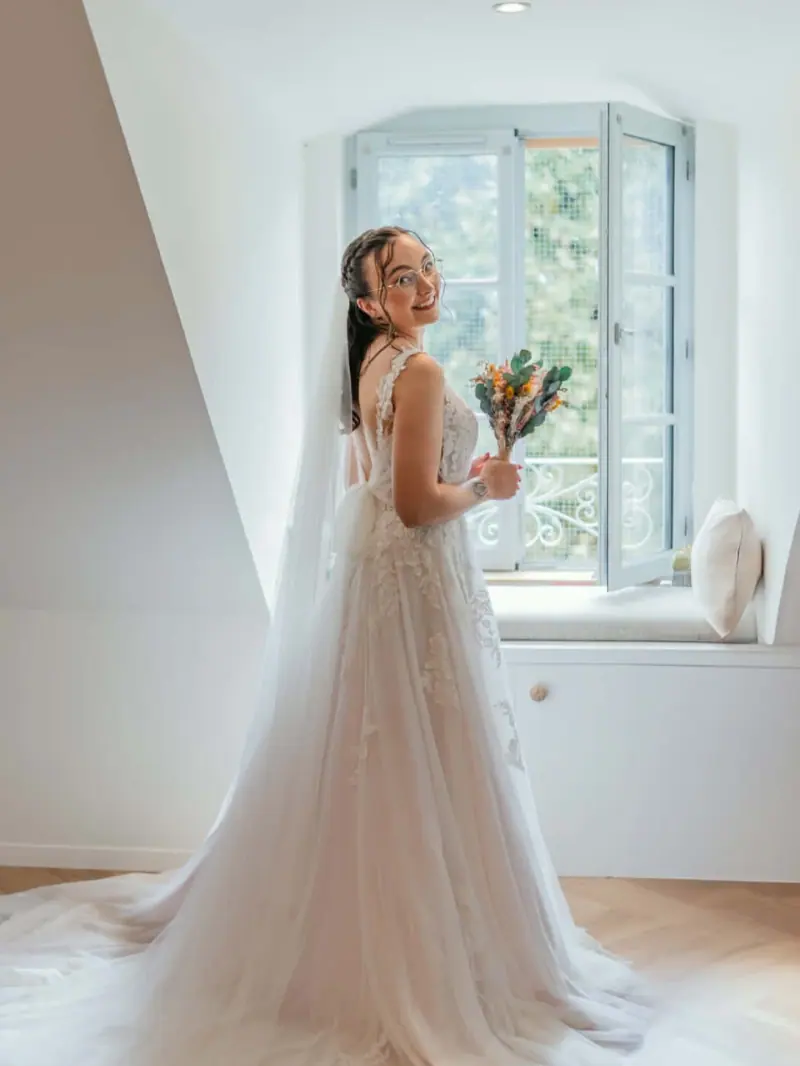 Mathilde en robe de mariée près d'une fenêtre, cheveux attachés et bouquet.